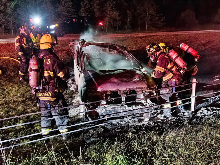 Firefighters in full gear extinguish a smoking, burnt-out car on the side of a road at night. The car is heavily damaged, surrounded by foam, with emergency lights illuminating the scene.