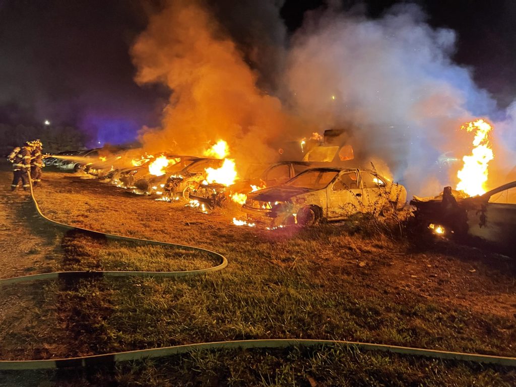 Several cars are engulfed in flames at night, with thick smoke rising into the sky. A firefighter is spraying water from a hose in an effort to control the blaze. The ground is illuminated by the fire.
