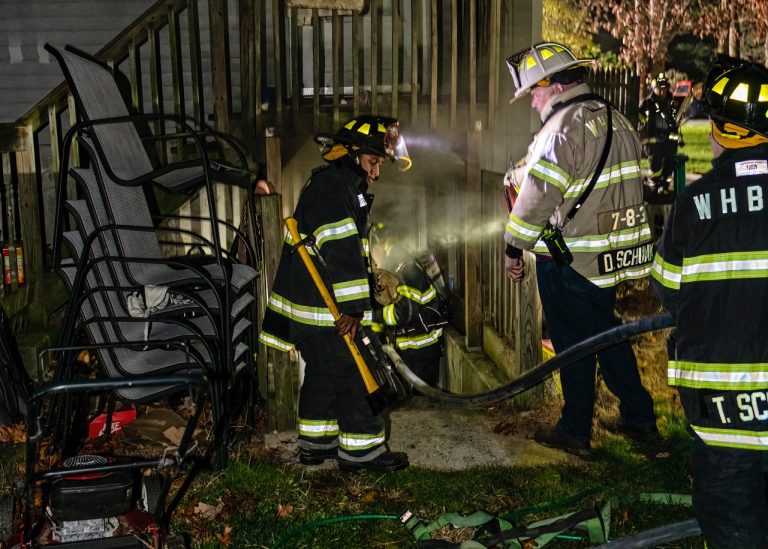 Three firefighters in gear work at the entrance of a basement at night. One holds an axe, another uses a flashlight, and the third oversees while holding a hose. Patio chairs and a lawnmower are nearby.