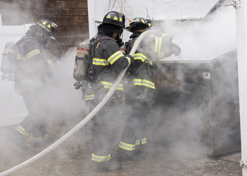 Three firefighters in full gear spray water into a smoking dumpster, surrounded by heavy white smoke, as part of an emergency response or training exercise outdoors.