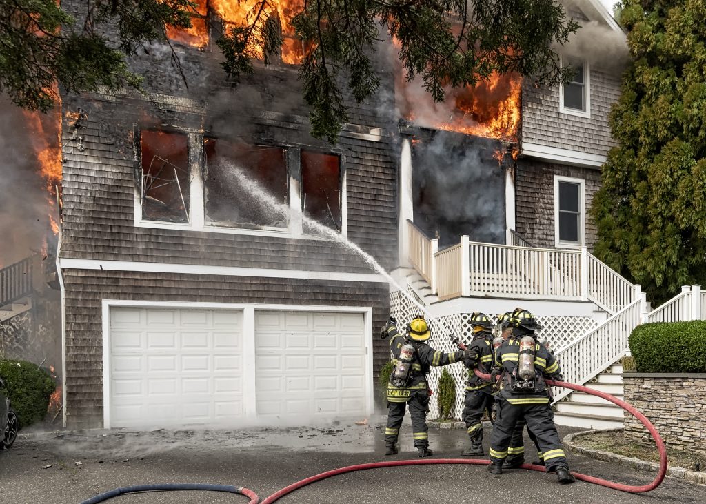 Four firefighters spray water at intense flames engulfing the upper floor of a two-story house, while thick smoke billows out the windows. The lower garage level is undamaged, and a staircase is visible on the right.