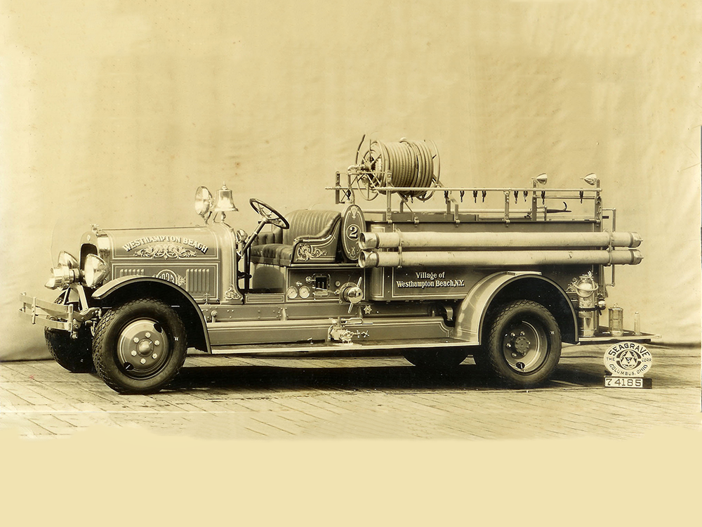 A vintage fire truck with a hose reel and firefighting equipment is parked on a brick surface, displaying "Village of Washington Board N.V.F.D." on its side. The photo has a sepia tone and a neutral backdrop.