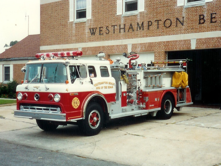 A vintage red and white fire truck is parked in front of a brick fire station with "Westhampton Beach" written above the garage doors. The truck has various firefighting equipment and a yellow hose on its side.