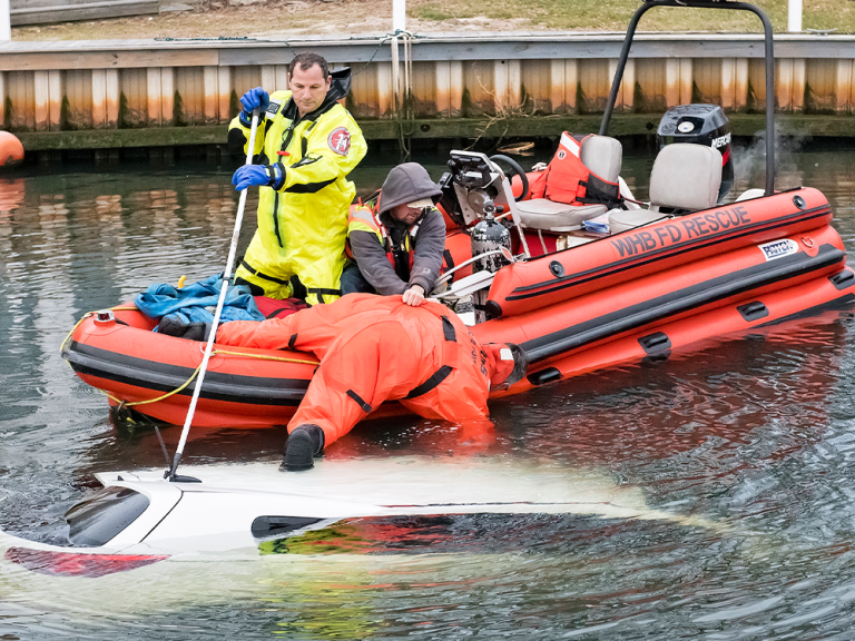 Two rescue workers in a red inflatable boat assist with the recovery of a partially submerged white car in the water near a dock. One worker leans over the boat, reaching toward the car.