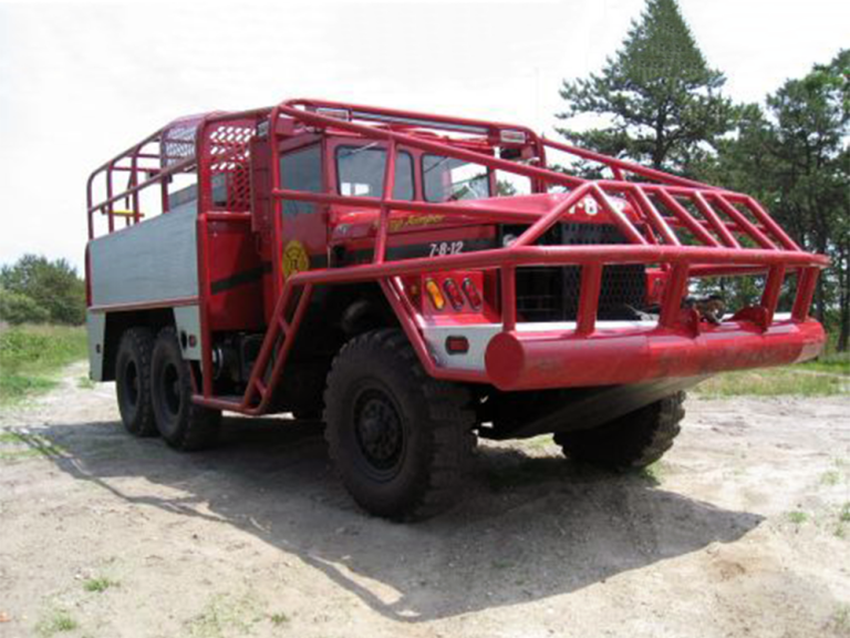 A large red off-road fire truck with a protective metal frame and six wheels is parked on a dirt surface outdoors, with trees visible in the background.