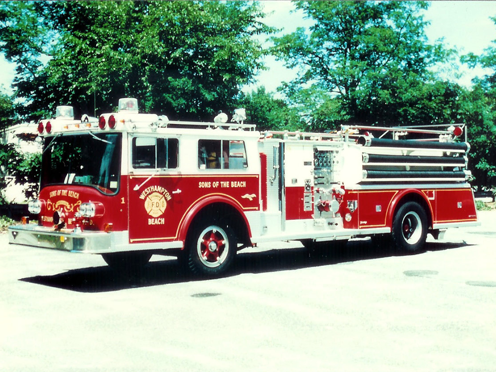 A red and white fire truck labeled "Sons of the Beach" is parked outdoors on a sunny day, surrounded by green trees and clear sky.