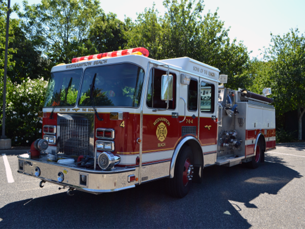 A red and white fire truck with lights on top is parked on a street in front of trees on a sunny day. The truck displays "Fire Department" markings and equipment along its side.