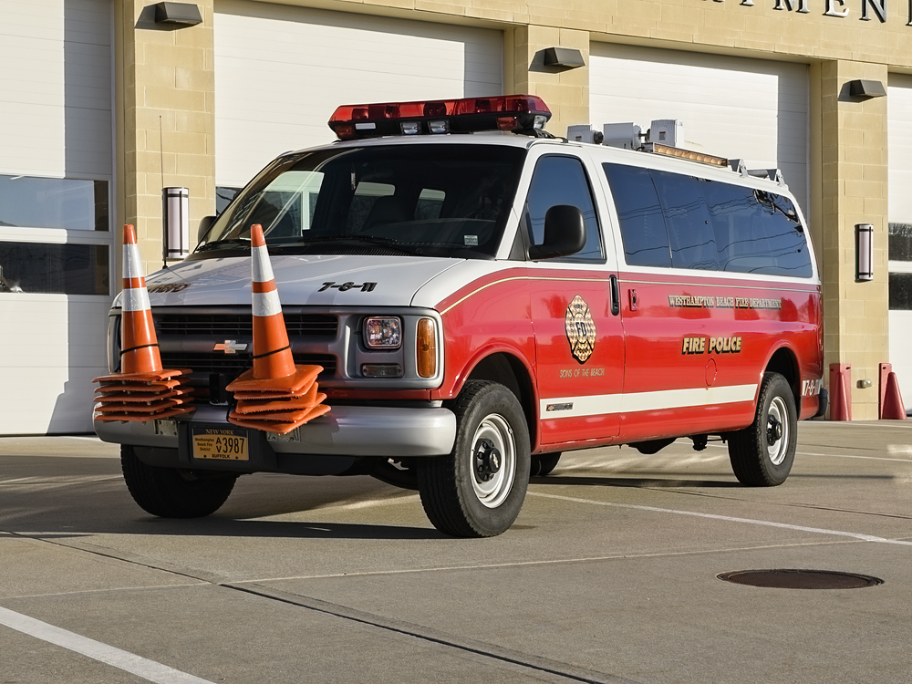 A red and white fire police van is parked in front of a fire station. Orange traffic cones are attached to the vehicle’s front bumper. The van has emergency lights on top and official markings on the side.