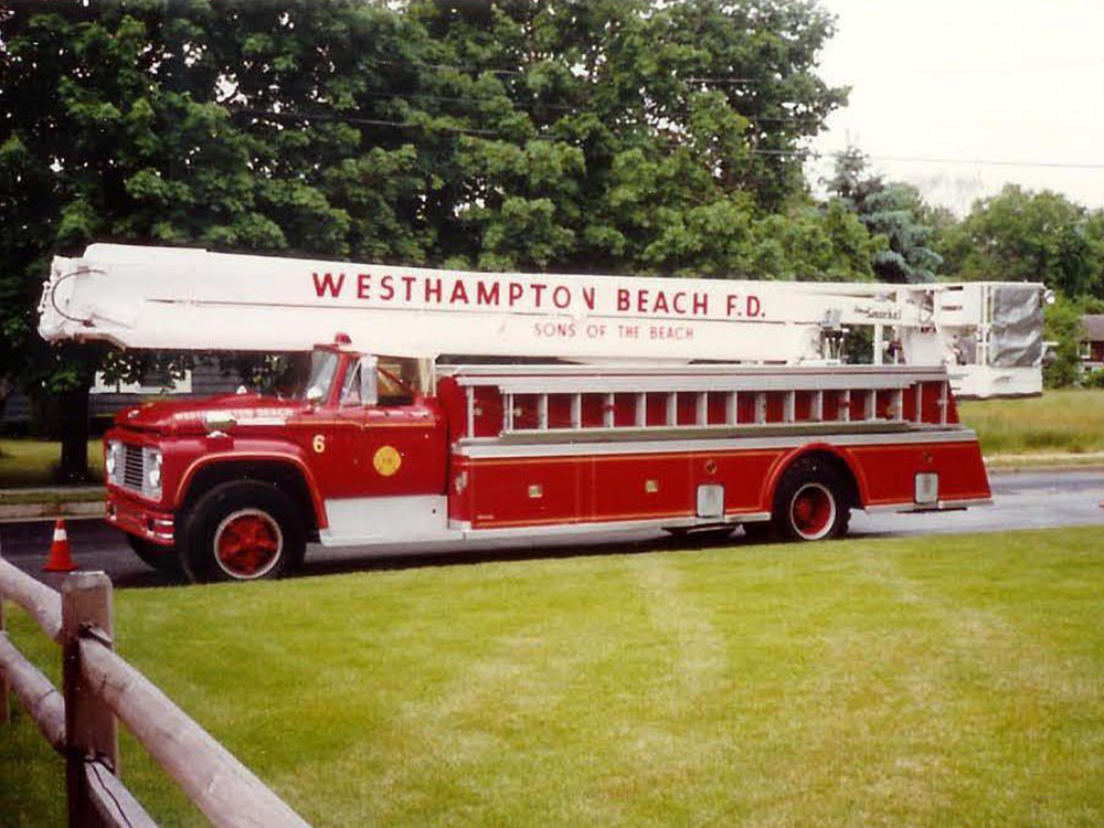 A red and white fire truck labeled "Westhampton Beach F.D. Sons of the Beach" is parked on a street beside a grassy area, with trees in the background.