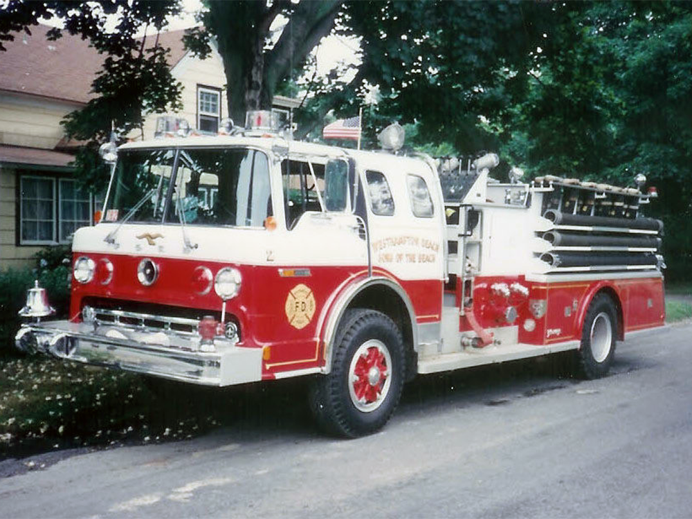 A vintage red and white fire truck is parked on a street beside trees and a house. The truck features chrome details, fire hoses, and fire department markings on the side.