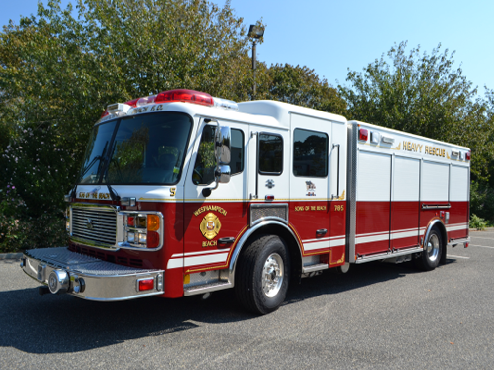 A red and white fire rescue truck with emergency lights parked on a paved road, surrounded by trees and greenery in the background. "Heavy Rescue" is written on the side of the truck.