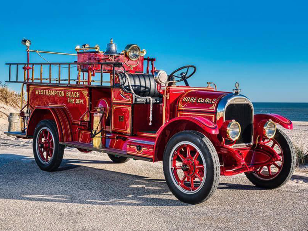 A vintage red fire truck with gold detailing, labeled "Westhampton Beach Fire Dept." and "Hose Co. No. 2," is parked on a sandy beach under a clear blue sky.