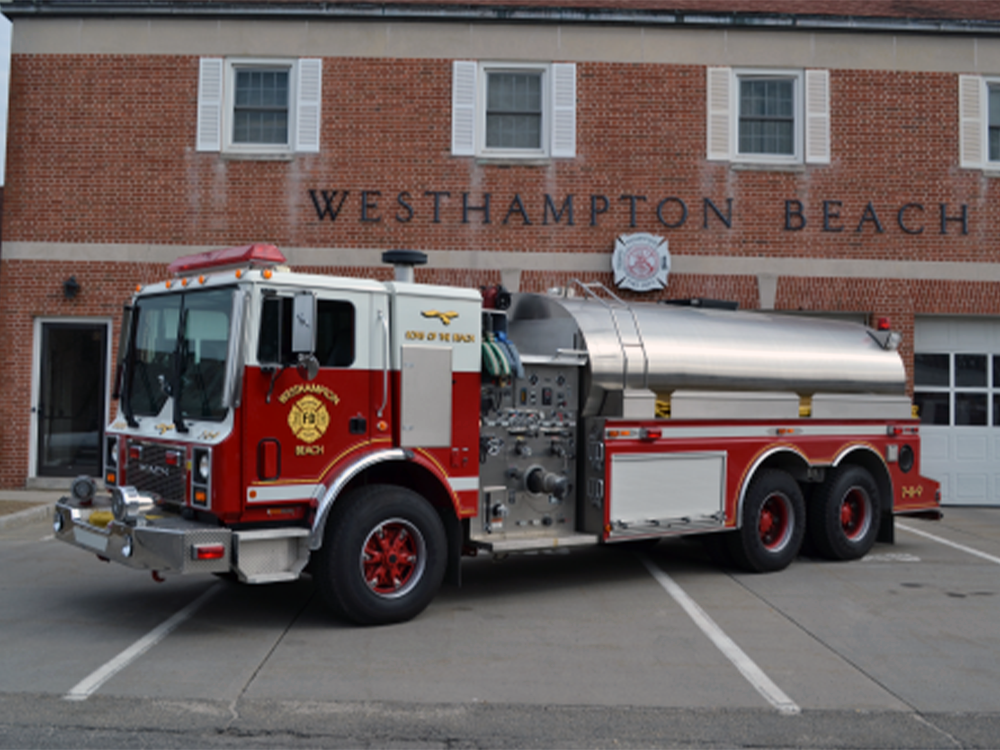 A red and white fire truck labeled "Westhampton Beach" is parked outside a brick fire station with "WESTHAMPTON BEACH" written above the entrance.