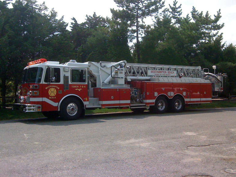A red fire truck with a long ladder labeled "Westhampton Beach" is parked on a paved road next to green trees on a sunny day.