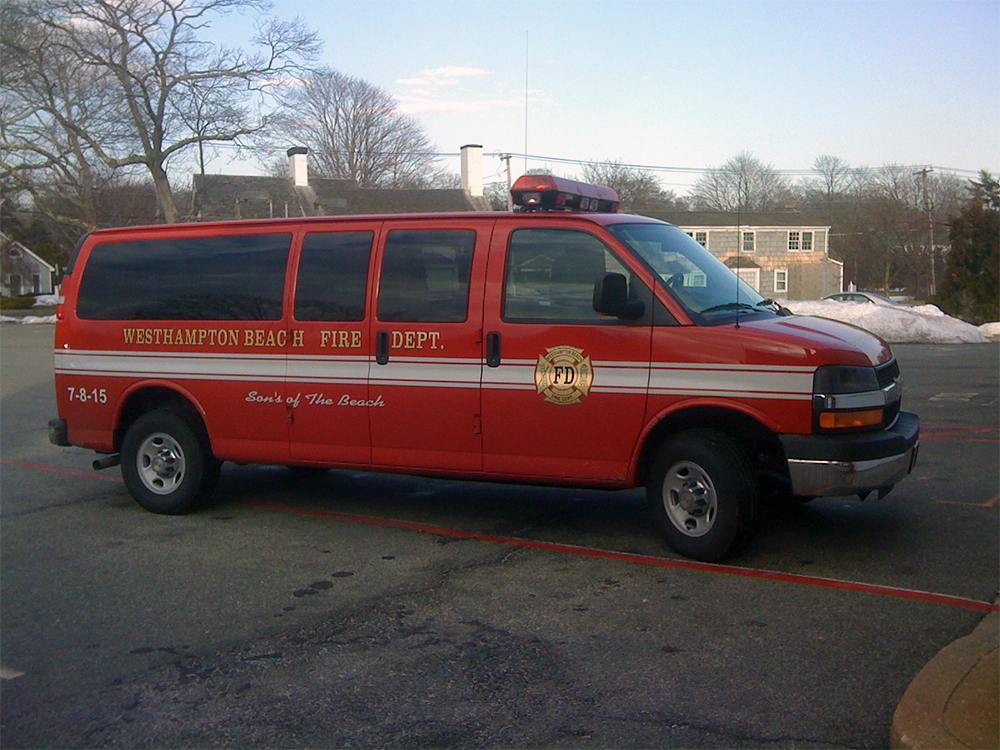 A red Westhampton Beach Fire Department van with emergency lights is parked in a lot near residential buildings on a clear day, with some snow visible on the ground in the background.