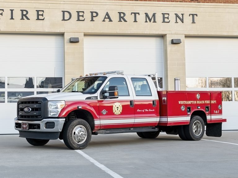 A red and white fire department truck is parked in front of the Westhampton Beach Fire Department building with its garage doors closed. The truck has emergency lights and fire department logos on the door and side.