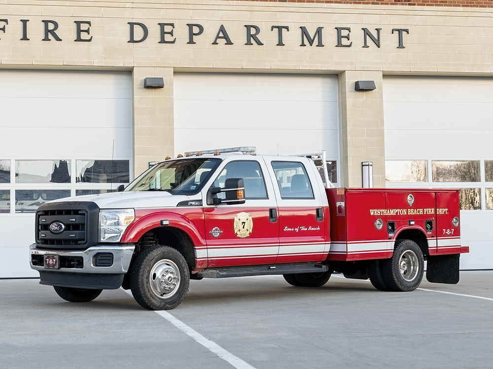 A red and white fire department truck is parked in front of the Westhampton Beach Fire Department building with its garage doors closed. The truck has emergency lights and fire department logos on the door and side.