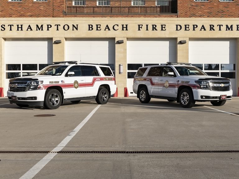 Two white fire department SUVs with red stripes parked in front of the Westhampton Beach Fire Department building, which has large white garage doors and a brick facade.